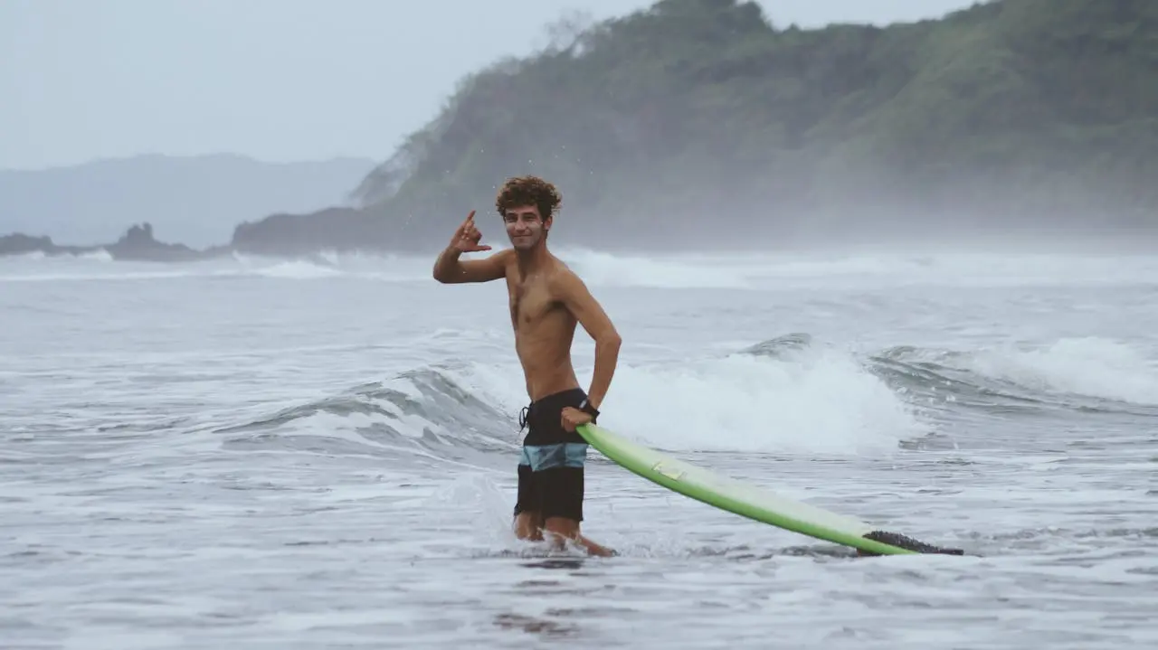 Beginner surfer standing on a longboard at Jacó Beach, Costa Rica, with green hills behind