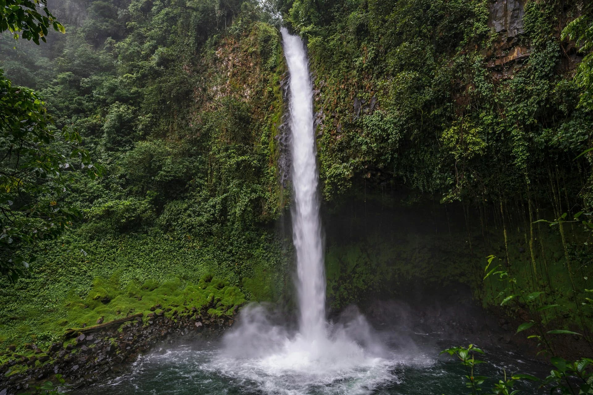 Rain-soaked green rainforest and a swollen waterfall near La Fortuna, Costa Rica, with the Arenal Volcano hidden in low cloud